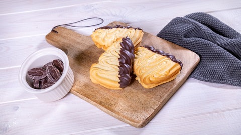 Drei Gebäckstücke in Wellenform mit dunkler Kuvertüre an einer Seite liegen auf einem Brett, daneben ein weißes Schälchen mit runden dunkelbraunen Schokostücken für das Flammende Herz und auf der rechten Seite ein graues Geschirrtuch