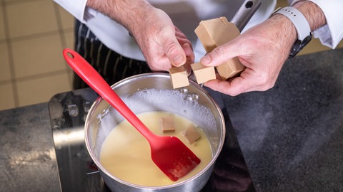 Eine Hand hält hellen Nougat für den Nachtisch, die andere schneidet ihn in einen Topf, der auf einem Herd steht und die Stücke fallen in eine gelbe Flüssigkeit, die über das Lebkuchenparfait soll