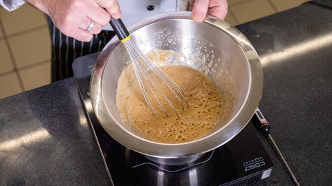 Auf einer Herdplatte steht ein Topf mit einer Metallschüssel obendrauf, eine Hand hält die Schüssel in der anderen ist ein Schneebesen, mit dem eine bräunliche Flüssigkeit für den Nachtisch das Lebkuchenparfait aufgeschlagen wird
