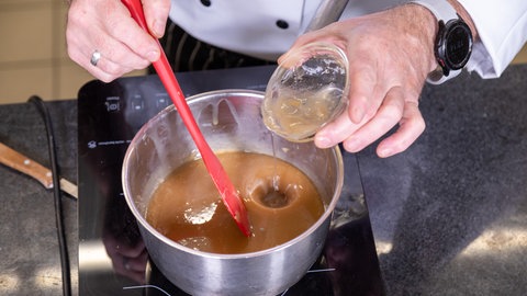 Eine Hand mit rotem Kochlöffel rührt in einem Topf, der auf dem Herd steht und in dem sich für den Nachtisch eine bräunliche Flüssigkeit befindet. Die andere Hand hält eine kleine Glasschale, in der sich eine helle Flüssigkeit befindet.