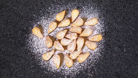 Die fertigen, abgekühlten Mutzenmandeln liegen mit Puderzucker bestäubt, wie im Rezept vorgeschlagen, auf einer schwarzen Oberfläche.