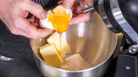 Ein Ei wird über einer Schüssel aufgeschlagen, in dem sich Butter und Zucker wie im Rezept beschrieben für den Teig der Mutzenmandeln befindet.