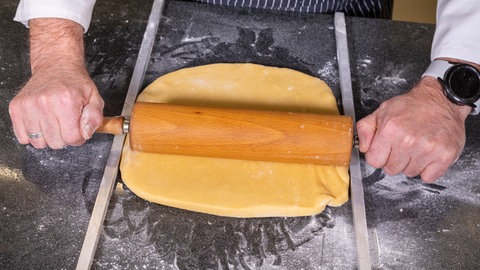 Zwischen zwei Metallstäben wird mit einem Nudelholz der Teig für die Mutzenmandeln ausgerollt, damit gleich die Form zum Ausstechen eingesetzt werden kann.
