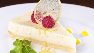 Rezept Zitronentarte mit Himbeeren: Ein Stück Zitronentarte liegt auf einem weißen Teller, garniert mit Minzeblättern