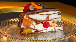 Ein Stück der SWR1 Pfännle Schoko-Flädle-Torte mit Früchten steht auf einem Glasteller. Die Torte ist in Schichten aufgebaut, die weiße Creme ist mit verschiedenen frischen Früchten gefüllt. Oben eine dicke Schicht Schokoglasur, dekoriert mit Trauben, Himbeeren, Blaubeeren und Aprikosen.