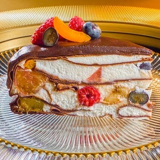 Ein Stück der SWR1 Pfännle Schoko-Flädle-Torte mit Früchten steht auf einem Glasteller. Die Torte ist in Schichten aufgebaut, die weiße Creme ist mit verschiedenen frischen Früchten gefüllt. Oben eine dicke Schicht Schokoglasur, dekoriert mit Trauben, Himbeeren, Blaubeeren und Aprikosen.
