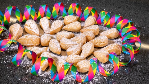 Fertige Mutzenmandeln liegen mit Puderzucker bestäubt, wie im Rezept vorgeschlagen, auf dunklem Untergrund umringt von einer bunten Luftschlange.