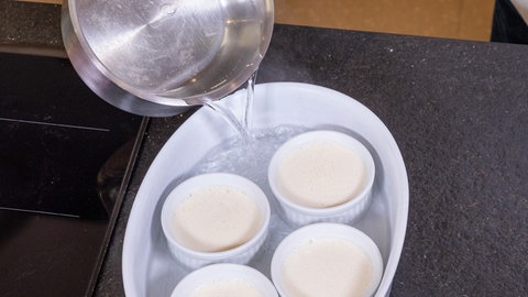 Eine Aufflaufform wird mit heißem Wasser befüllt. In der Form stehen kleinere Förmchen mit der Crème Brûlée.