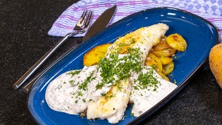 Neben einem blauen, rechteckigen Teller liegt Fischbesteck. Auf dem Teller das Osteressen: ein Stück Zanderfilet mit Zitronenabrieb bestreut liegt auf Röstkartoffeln und alles ist mit einer weißen Soße übergossen und mit Dill bestreut. Ein passendes Rezept für Ostern.