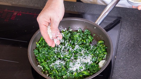 Fisch in Mandelkruste mit Spinat: In einer Pfanne wird Parmesan dem Blattspinat zugefügt.