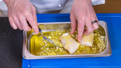 Ein helles Fischfilet vom Kabeljau wird in Olivenöl und Zwiebeln eingelegt und gewendet, damit es dann in den Ofen und später in der Sauce schwimmen kann.