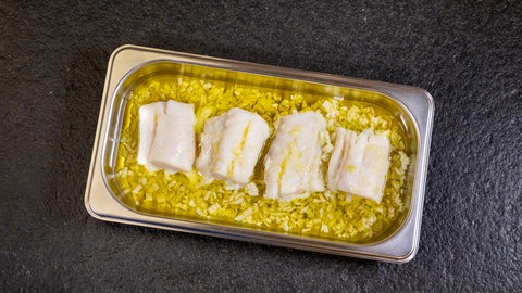 Vier helle Fischfiletstücke vom Kabeljau liegen in einer rechteckigen Metallschüssel in einer Ölmarinade. Sie wurden im Ofen gegart und werden später mit Sauce übergossen.