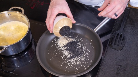 Helle kleine Samen liegen in einer Pfanne auf dem Herd, zu denen schwarze Samen dazugeschüttet werden. Der Sesam wird für den Reis geröstet und dann zum Kabeljau und der Sauce gereicht.