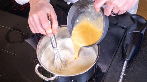 Eine gelbe Flüssigkeit, die Eier mit der Sahne, wird aus einer Schüssel in eine weiße Flüssigkeit – die Sauce für den Kabeljau und Reis – in einen Topf gegossen. 