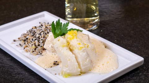 Kabeljau aus dem Ofen mit Sauce und Reis: Ein helles Fischfilet liegt auf Reis mit Sesamkörnern und ist mit einer hellen Sauce übergossen. Das Ganze ist schön auf einem weißen rechteckigen Teller angerichtet.