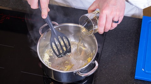 Eine durchsichtige, gelbliche Flüssigkeit – Weißwein – wird in den Topf mit angebratenen Zwiebeln aus einem durchsichtigen Messbecher gegossen. Daraus wird die Sauce für den Kabeljau und Reis.