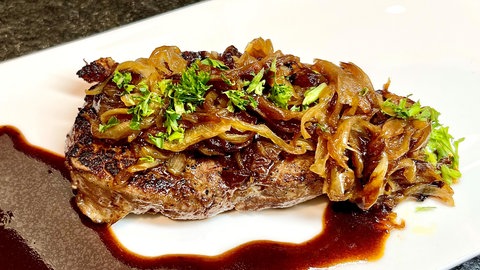 Saftiger Zwiebelrostbraten. Das Rind-Fleisch istz auf einem Teller mit geschmelzten Zwiebeln und Soße angerichtet ist.
