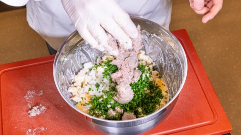 In einer Schüssel mit der Knödelmasse liegt klein geschnittener Schnittlauch oben auf und eine Hand mit weißem Gummihandschuh gibt die Pfälzer Leberwurst hinzu.