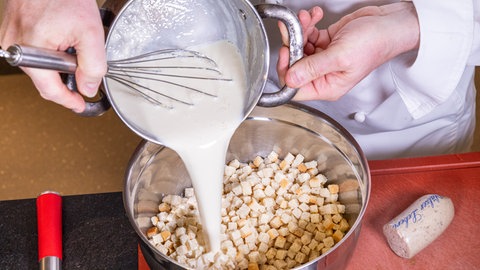 Erwärmte Milch wird aus einem kleinen Topf in Schale über kleingeschnittene Brötchen gegossen. Daneben liegt eine halbe Rolle Pfälzer Leberwurst.