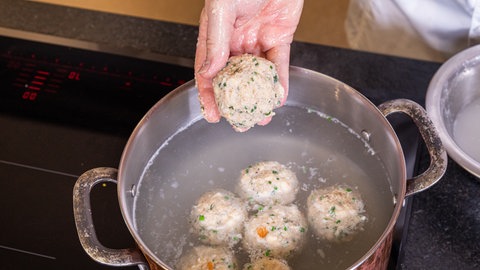 Eine Hand lässt die Knödel mit Pfälzer Leberwurst in einen Topf mit heißem Wasser gleiten, der auf einem Herd steht.