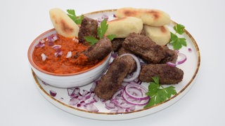 Ćevapčići mit Pizzabrotsticks, Ajvar, Zwiebeln und Petersilie auf einem Teller. Ein echter Grill-Klassiker von SWR1 Pfännle-Koch Eberhard Braun: herzhafte CevapciciĆevapčići mit knusprigen Pizzabrotsticks zum Snacken während der Fußball-EM-Spiele.