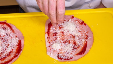Eine mit Tomatenmark bestrichene Schinkenscheibe wird mit Käse bestreut. 