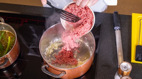 In einem Topf, in dem schon Zwiebeln braten, wird Hackfleisch aus einer Schüssel mit einem Pfannenwender hinzugefügt.