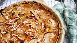 Apfeltarte mit Mandelstiften in einer runden Backform, daneben grünkarierte Kochhandschuhe