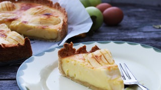 Stück Elsässer Apfelkuchen auf einem Teller, Äpfel und ein angeschnittener Kuchen im Hintergrund