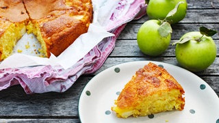 Stück Rührteigkuchen mit Äpfeln auf Teller, daneben ein angeschnittener Kuchen in Backform und Äpfel