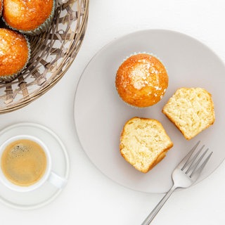 Auf einem Teller sind zwei Zitronenmuffins, einer von beiden ist halbiert. Daneben stehen eine Tasse Kaffee und ein Korb mit weiteren Muffins.