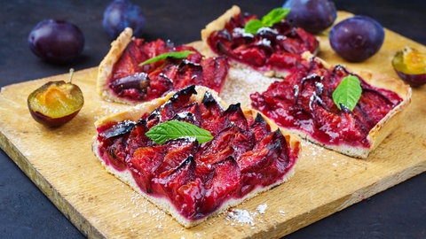 Rezept für Zwetschgenkuchen: Stück Zwetschgenkuchen mit Minzblatt als Deko auf Holzbrett, daneben ganze Früchte und dahinter ein ganzer Zwetschgenkuchen