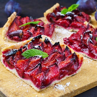 Rezept für Zwetschgenkuchen: Stück Zwetschgenkuchen mit Minzblatt als Deko auf Holzbrett, daneben ganze Früchte und dahinter ein ganzer Zwetschgenkuchen