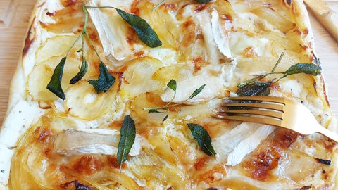 Zwiebelkuchen mit Blätterteig gebacken, darauf Ziegenkäse und Salbei. Der Kuchen liegt auf einem Holzbrett mit einer goldenen Gabel in der Mitte