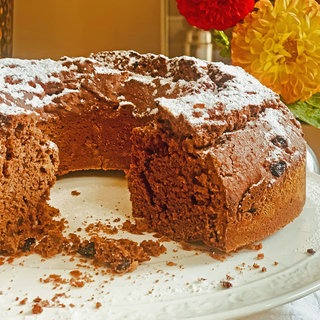 Rezept für Schokokuchen mit Mandeln und Rosinen: Ein angeschnittener Schokokuchen auf einer Tortenplatte, im Hintergrund orange-farbene Blüten.