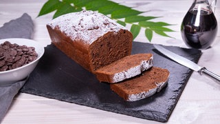 Rotweinkuchen auf einem schwarzen Schieferbrett, im Hintergrund steht eine Glaskaraffe mit Rotwein, links neben dem Kuchen ein weißes Schüsselchen mit Schokoraspeln und hinter dem Kuchen liegt ein grüner Zweig mit Blättern.