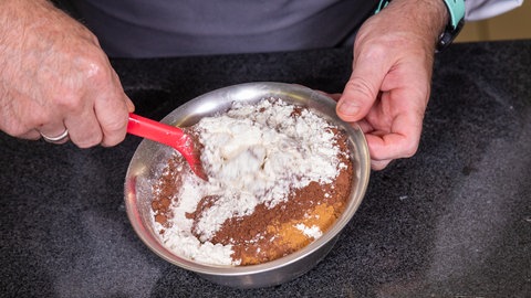 Ein Konditor mischt Backpulver, Zimt, Mehl und Kakao in einer kleinen Schüssel zu einer Masse.