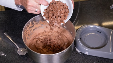 Ein Konditor gibt eine kleine Schüssel Schokoraspeln in eine Rührschüssel mit Teigmasse eines Rotweinkuchens.