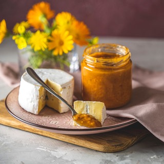Ein Teller mit einem angeschnittenen Camembert-Käse, einem geöffneten Glas Chutney aus Früchten wie Vogelbeeren, einem Löffel mit Chutney und einem Stück Käse
