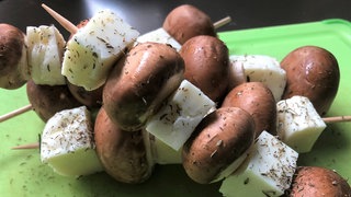 Zwei Champignon-Halloumi-Holzspieße auf einem grünem Schneidebrett. Pilze sind kalorienarm und machen lange satt. Vielfältig variiert sind sie ideal auf dem Grill oder als Antipasti. Gut gewürzt schmecken sie hervorragend als Ersatz für Fleisch.