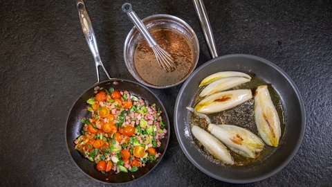 Chicoree gebraten: Eine Pfanne mit gebratenem Chicoree, ein Topf mit Dressing und eine Pfanne mit gebratenen Zwiebelwürfeln, Speckwürfeln und Kirschtomaten.