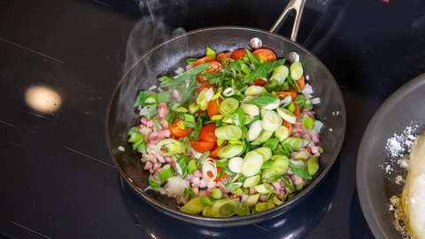 Für den gebratenen Chicoree wird ein Topping aus Speckwürfeln, Zwiebelwürfeln und Lauch vorbereitet und brät in einer Pfanne.