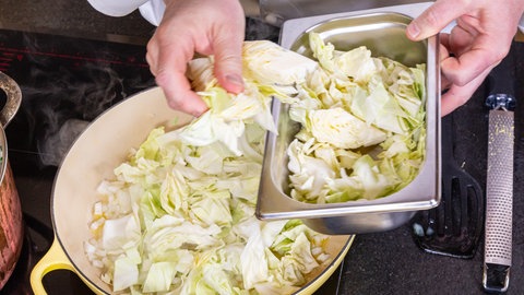 Eine Hand gibt klein geschnittenen Spitzkohl bzw. Filderkraut in eine Pfanne, die auf dem Herd steht.