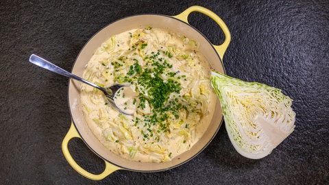 Ein Topf mit schwäbischem kleingeschnittenem Filderkraut in einer cremigen Soße mit einem Löffel drin. Daneben liegt ein aufgeschnittener halber Spitzkohl.