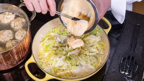 In einer Pfanne kocht der Spitzkohl bzw. das Filderkraut mit Pfeffer bestreut und eine Hand gibt aus einer kleinen Metallschale die Creme fraiche Eigelb Mischung mit einem Spatel in die Pfanne.