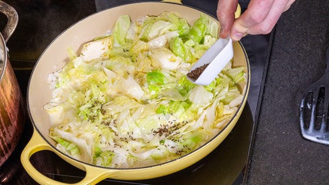 Eine Hand gibt aus einem kleinen weißen Schälchen ganzen Kümmel in die Pfanne mit dem Spitzkohl bzw. Filderkraut und der Sahne.