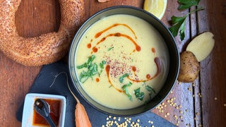 Linsensuppe in einer schwarzen Schale auf einem Holztisch. Drumherum liegen verschiedene Zutaten.