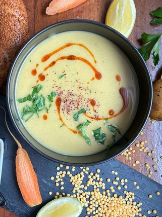 Linsensuppe in einer schwarzen Schale auf einem Holztisch. Drumherum liegen verschiedene Zutaten.