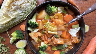 Auf einem Holztisch steht eine schwarze Schale mit Kohlsuppe. Um die Schale herum liegen Brokolli, Karotte, Zwiebel und Kohl.