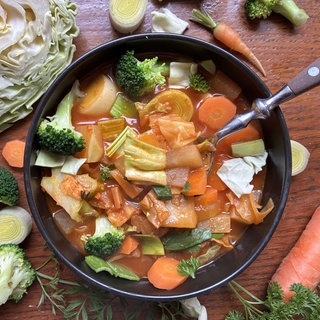 Auf einem Holztisch steht eine schwarze Schale mit Kohlsuppe. Um die Schale herum liegen Brokolli, Karotte, Zwiebel und Kohl.